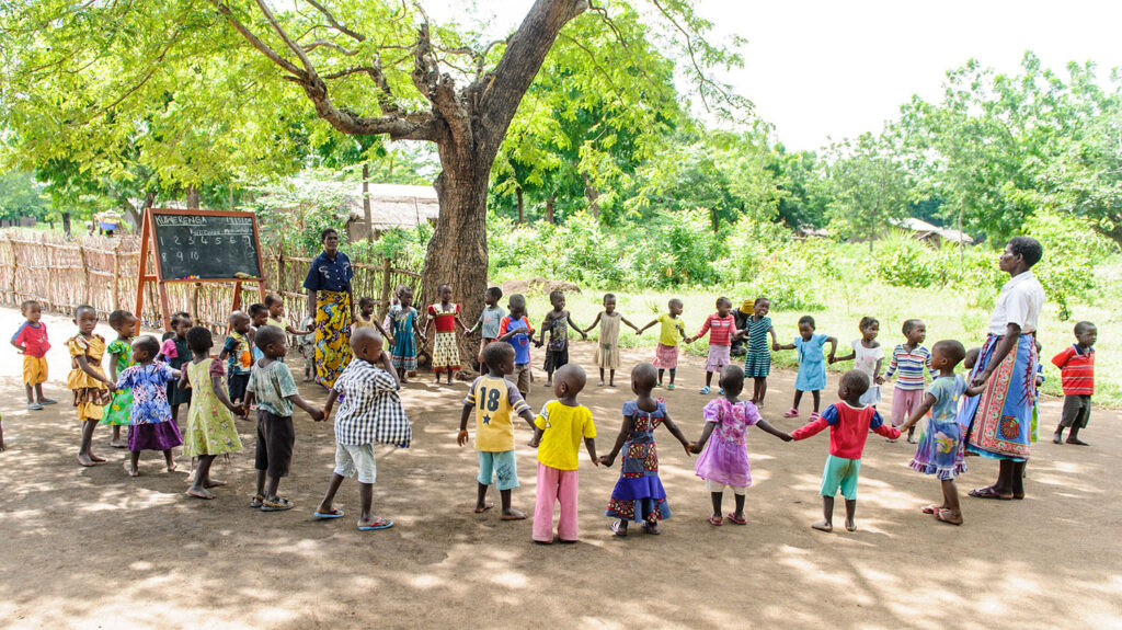 A large group of young children join hands to form a circle under the shade of a large tree.