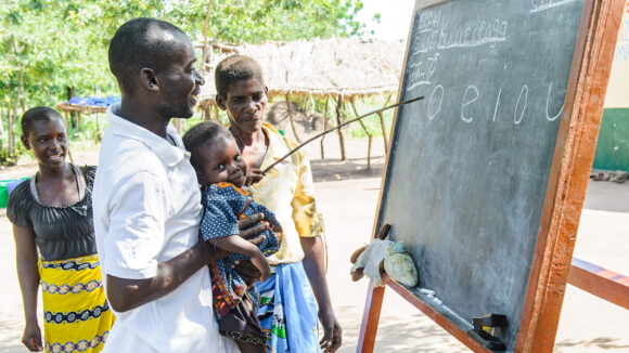 Outside, a male teacher stands in front of a blackboard holding a young child. In one hand he has a stick, which he is using to teach the letters on the blackboard. A man and woman also stand around the board, watching the lesson.