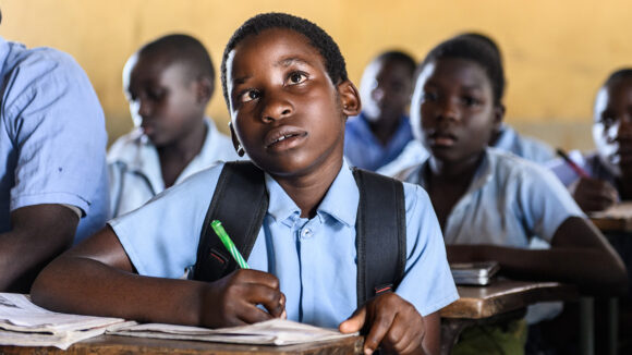 A young girl with a visual impairment sits writing at a desk in a classroom.
