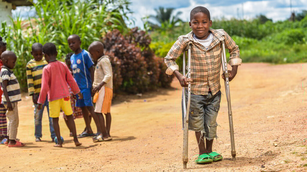 A boy stands smiling, assisted by crutches while a group of children gather behind him.