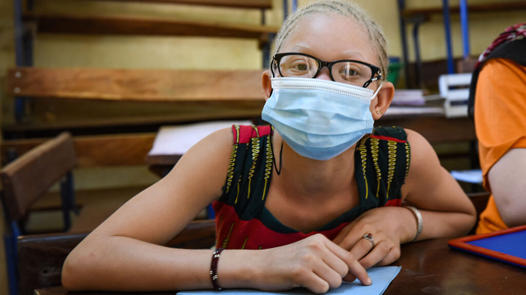 A girl, who has albinism and is wearing glasses and a mask, sits in a classroom.