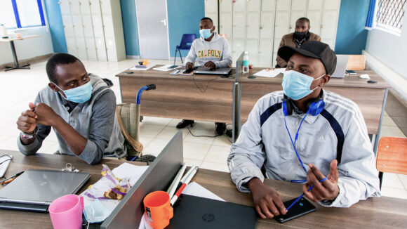 Two men sit at desks in a classroom while wearing face masks.