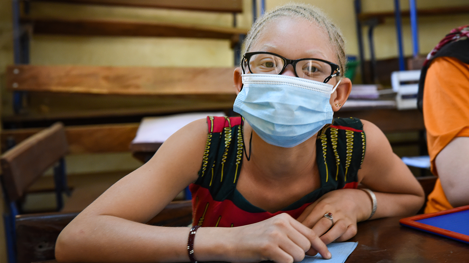 A young girl, who has albinism, sitting in a classroom. She is wearing a face mask.