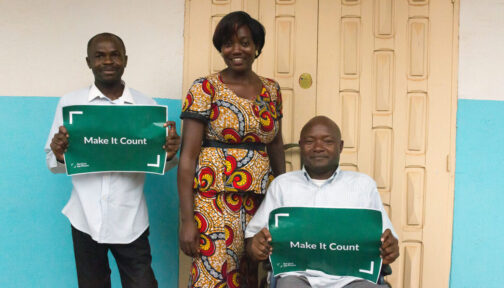 A woman and two men stand outside a building. The men, one of whom is sitting, are holding up signs with the words 'Make it count'.