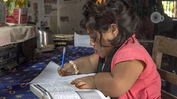 Reya from Bangladesh sits at a desk writing. She has shortened limbs and is wearing a pink blouse.