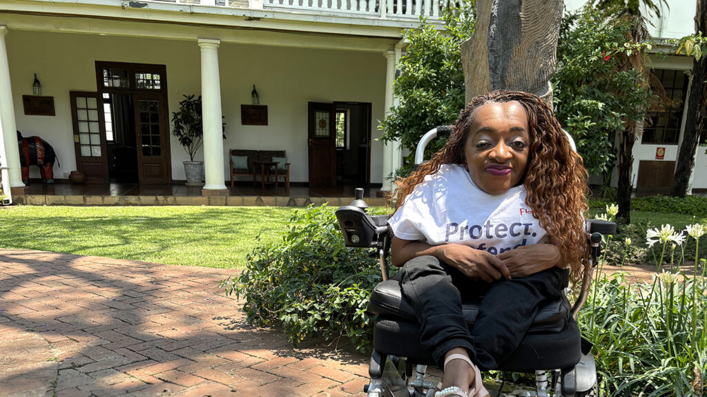 Florence wears a T-shirt with an Equal Zimbabwe logo. She's sitting in her wheelchair in a lush green garden.